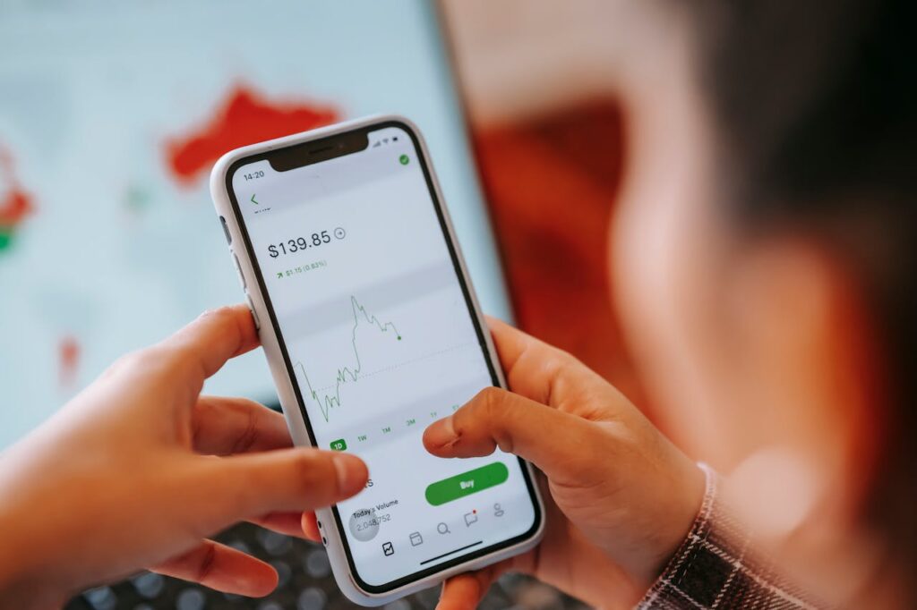 Bild einer Person, die mit einem Smartphone eine Börsen-App verwendet. Der Bildschirm des Handys zeigt einen Aktienkurs und einen "Buy"-Button, was auf eine Kaufoption hinweist