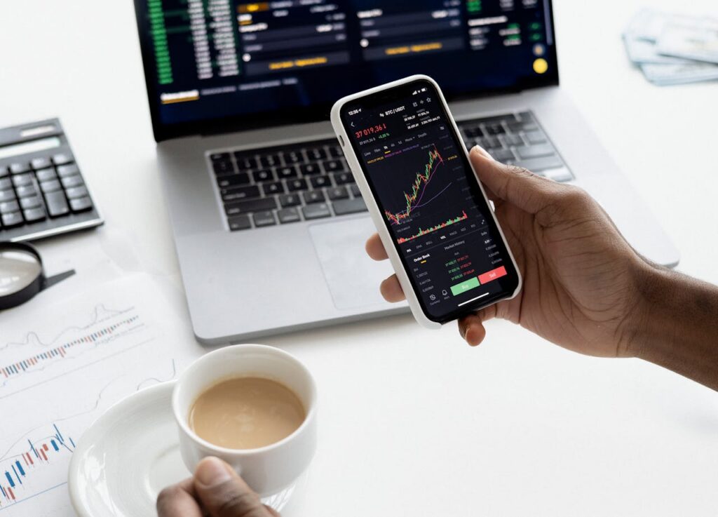 Portfolio aufbauen mit digitaler Unterstützung - Person analysiert Aktienkurse auf dem Smartphone – im Hintergrund Laptop, Finanzcharts und Kaffeetasse