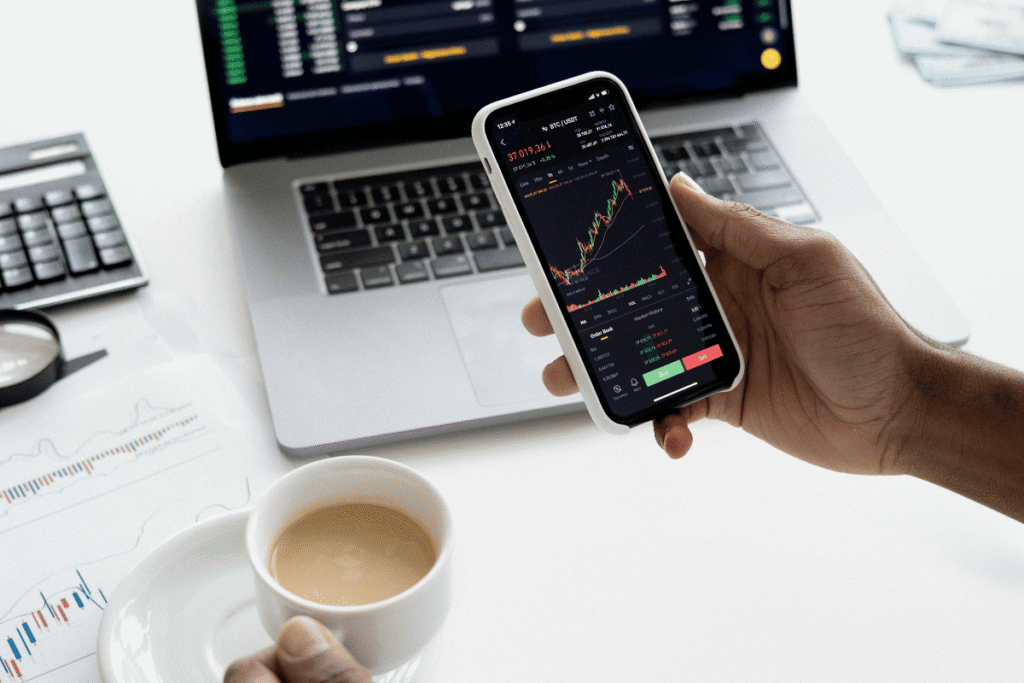 Portfolio aufbauen mit digitaler Unterstützung - Person analysiert Aktienkurse auf dem Smartphone – im Hintergrund Laptop, Finanzcharts und Kaffeetasse